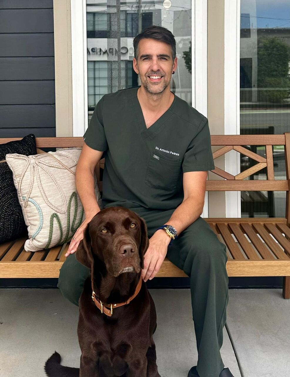 Dr. Antonio Pedraza sitting on a wooden bench with his chocolate Labrador