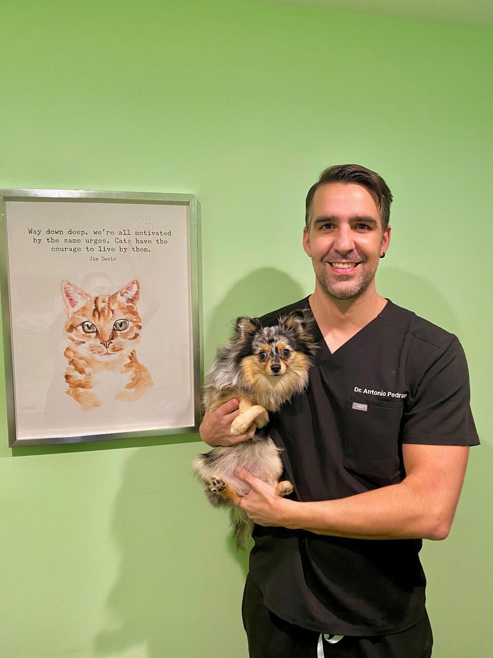 Dr. Pedraza holding a Pomeranian beside a framed cat watercolor